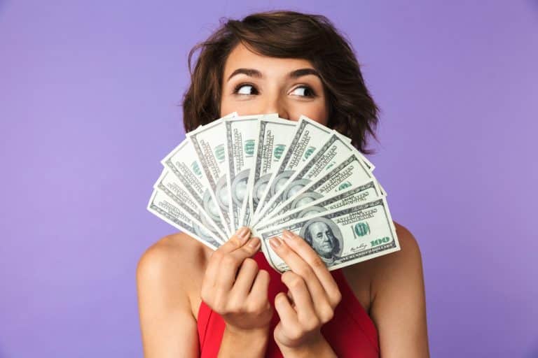 Brunette woman fanning a stack of 100 dollar bills in front of her face against a solid purple background. Human life value approach.